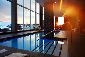 An indoor swimming pool with clear glass walls, a view of parked cars outside, and a sunset visible through the windows.
