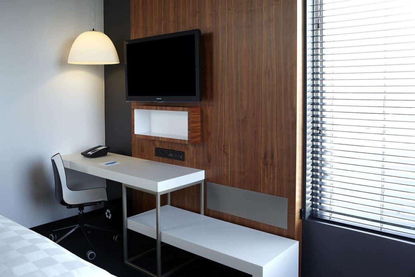 A hotel room with a desk, chair, television, and a window with blinds.