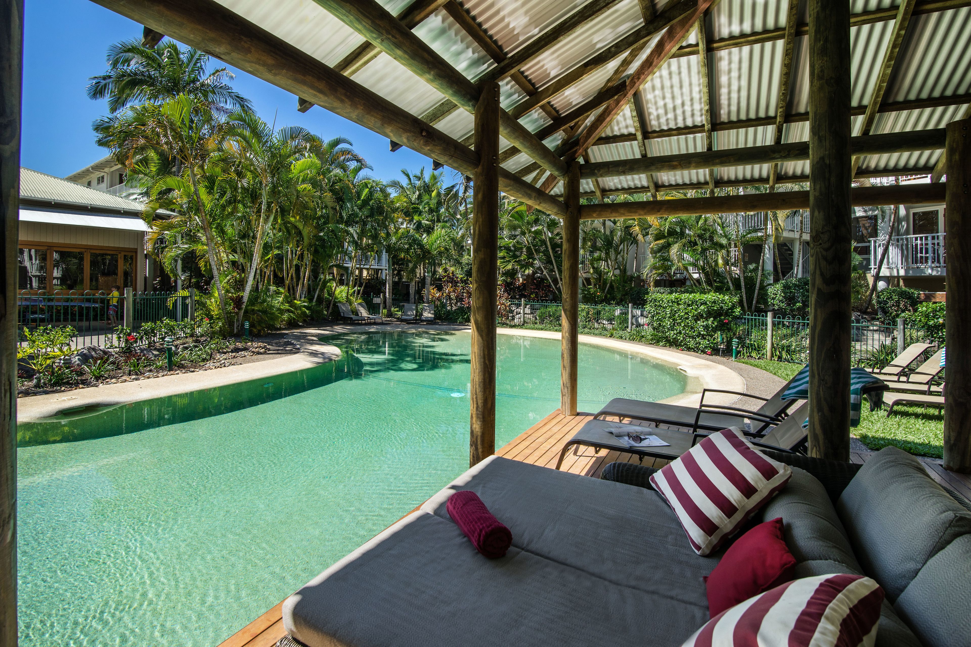 A poolside area with a covered seating area, a pool, and surrounding tropical vegetation.