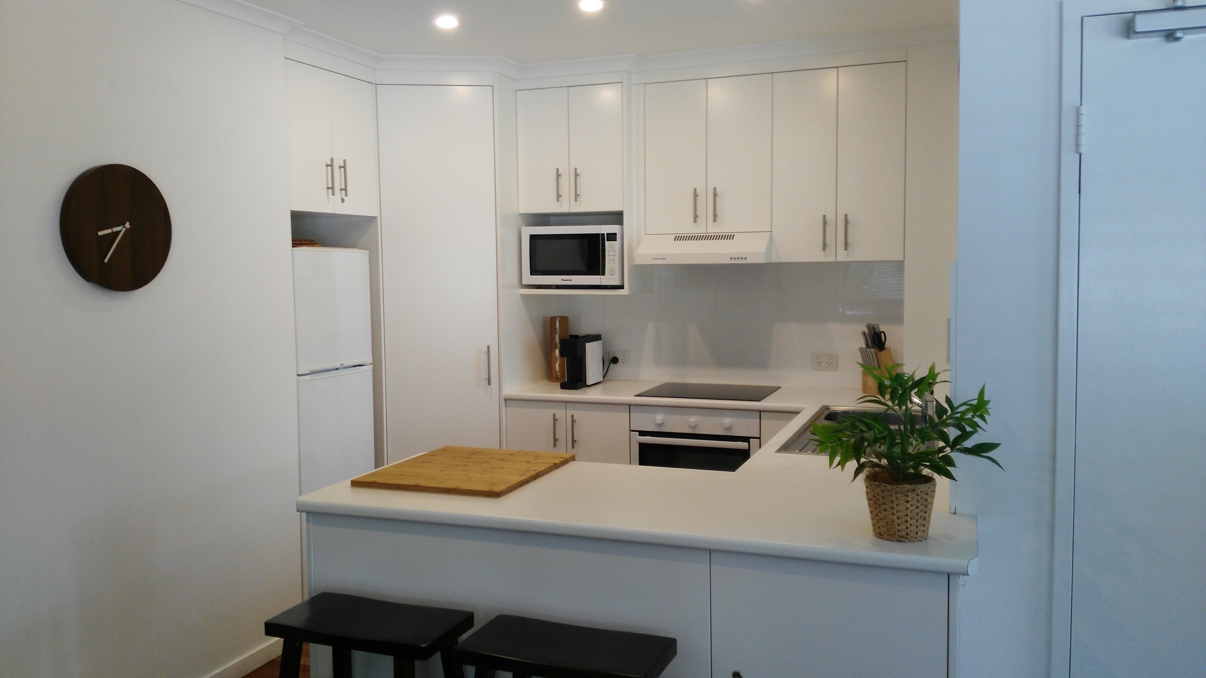 A compact kitchen with white cabinets, a microwave, a stove, and a sink. There is a clock on the wall and a plant on the counter.