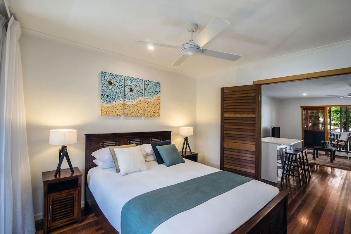 A hotel room with a bed, two bedside tables, a ceiling fan, and a view of the dining area.