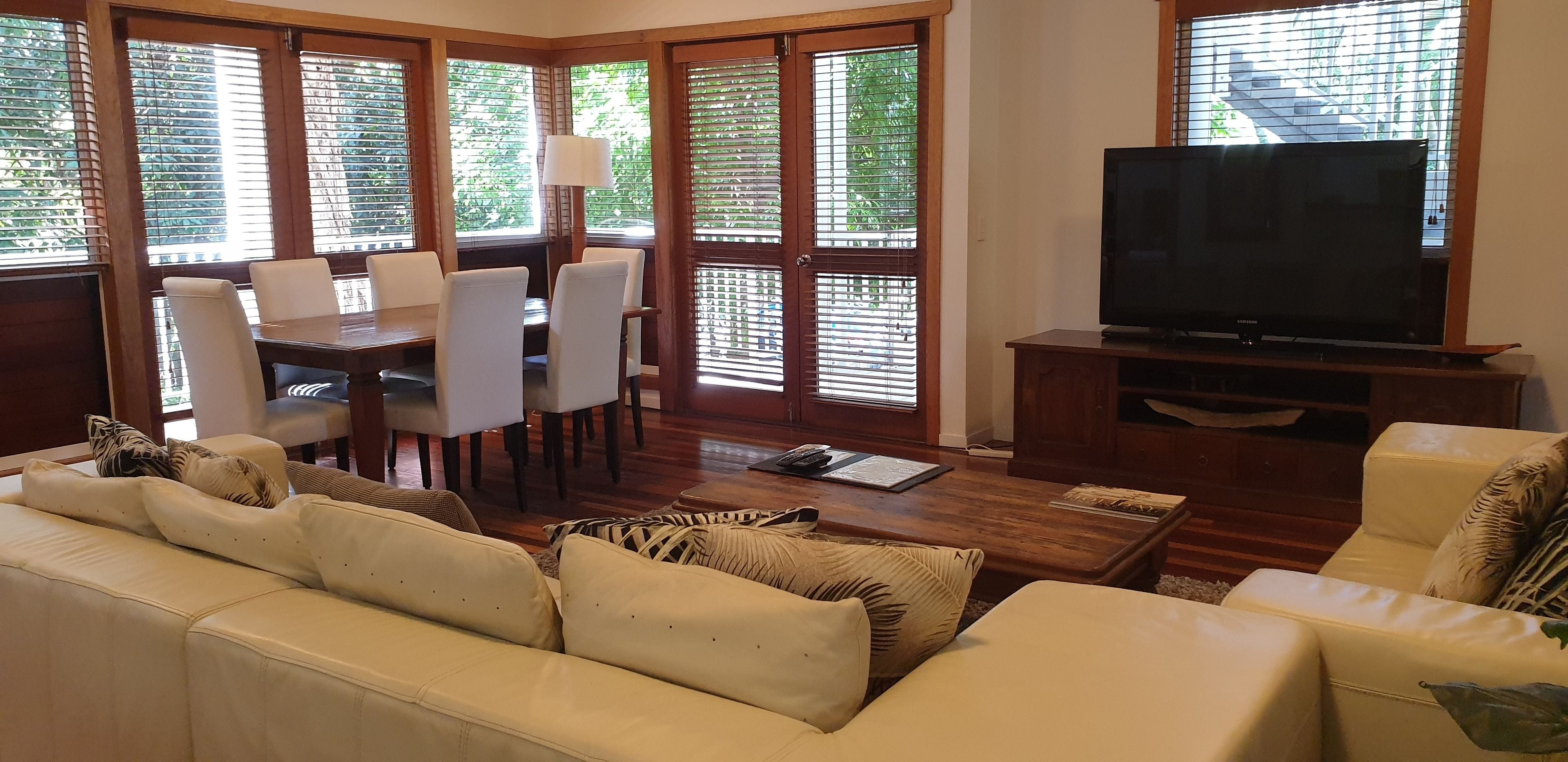A living room with a sofa, a dining table with chairs, a TV on a wooden stand, and large windows with blinds.