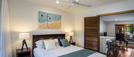 A hotel room with a large bed, two bedside tables with lamps, a ceiling fan, and a view into the dining area.