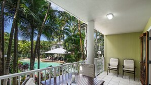 Un balcón con una mesa de madera, dos sillas y vistas a una piscina y palmeras.