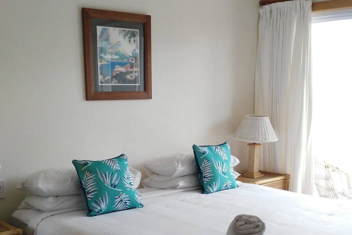 Una cama bien hecha con sábanas blancas y dos almohadas color turquesa, un cuadro en la pared, una lámpara de noche y una ventana con cortinas.
