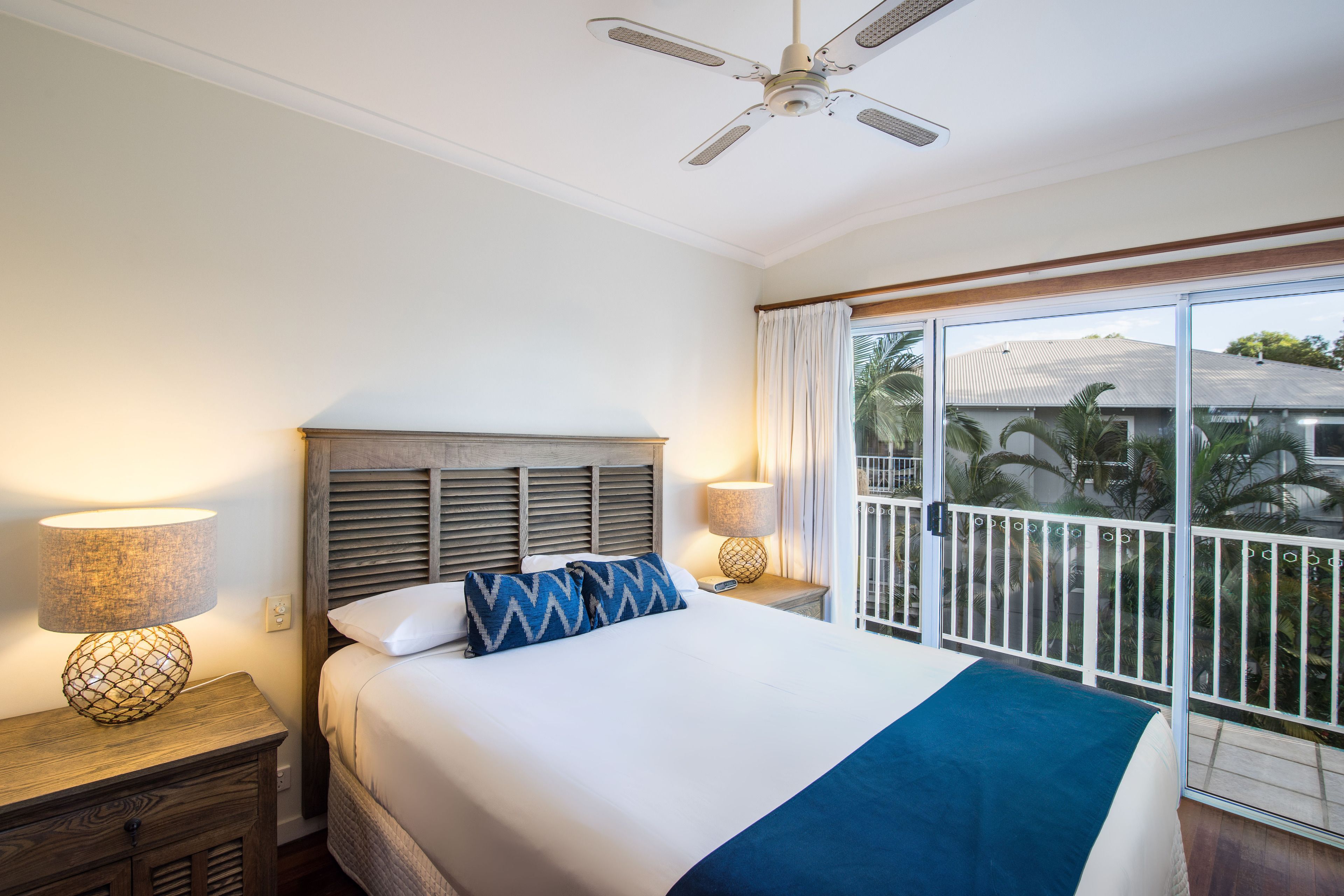 A hotel room with a bed, two bedside tables with lamps, a ceiling fan, and a balcony with a view of palm trees.