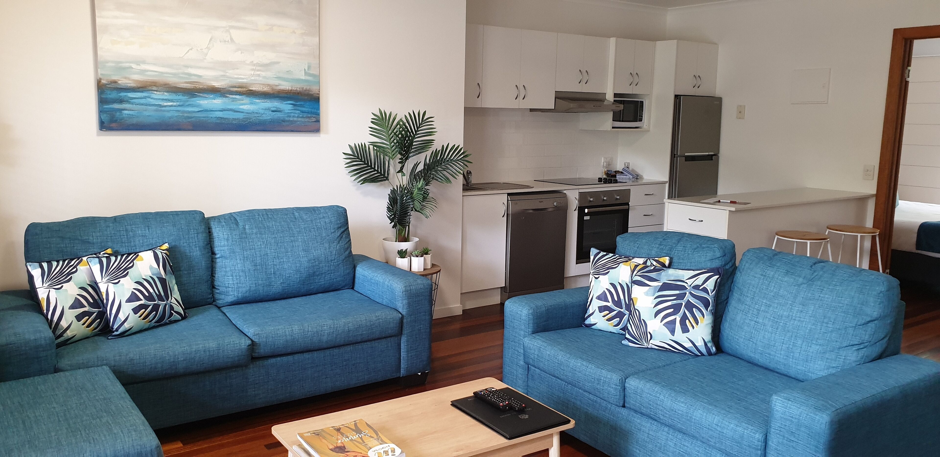 Una sala de estar con dos sofás azules, una mesa de centro y una cocina con armarios y electrodomésticos blancos.