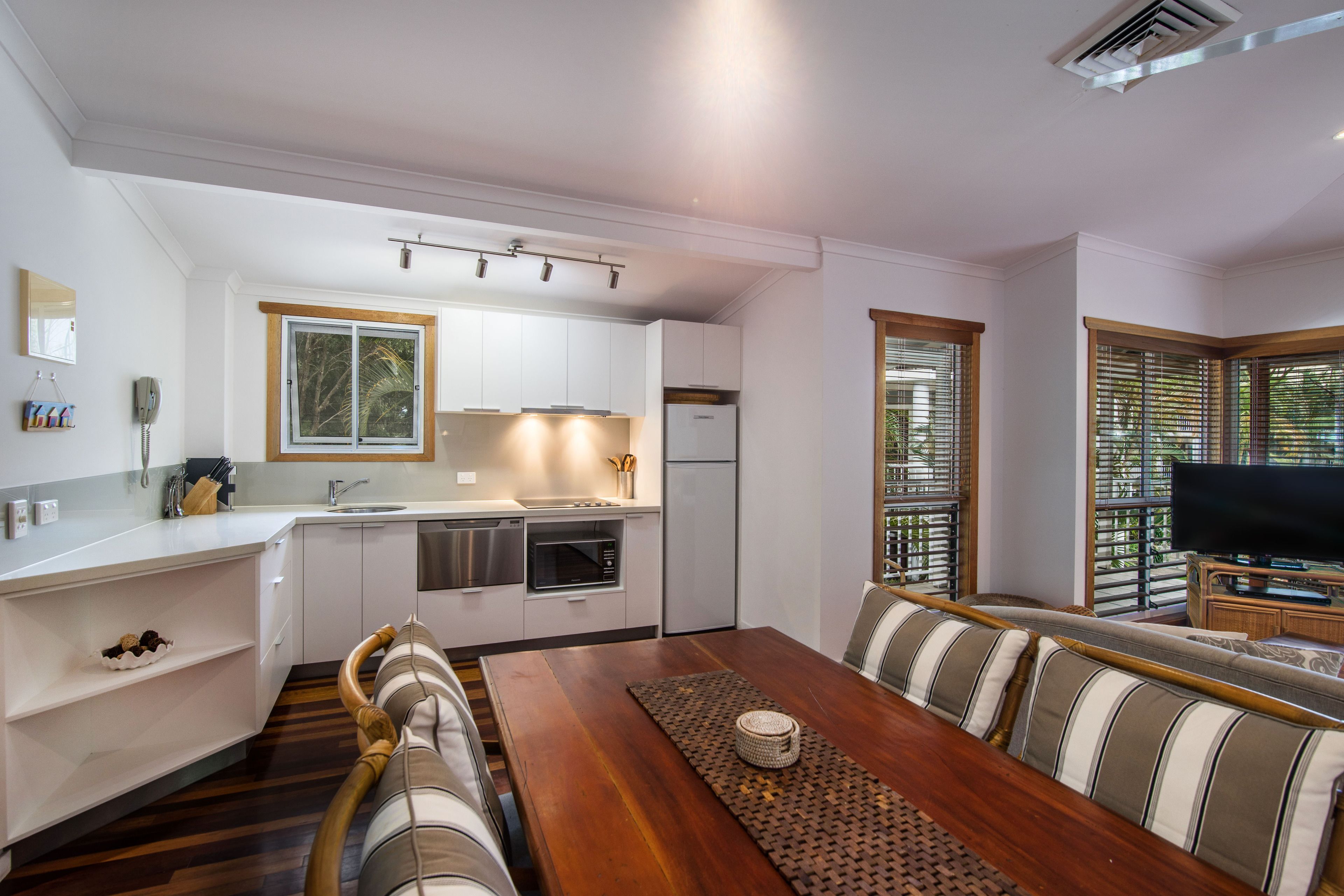 A modern kitchen with white cabinets, a dining area with a wooden table and striped cushions, and a TV in the corner.