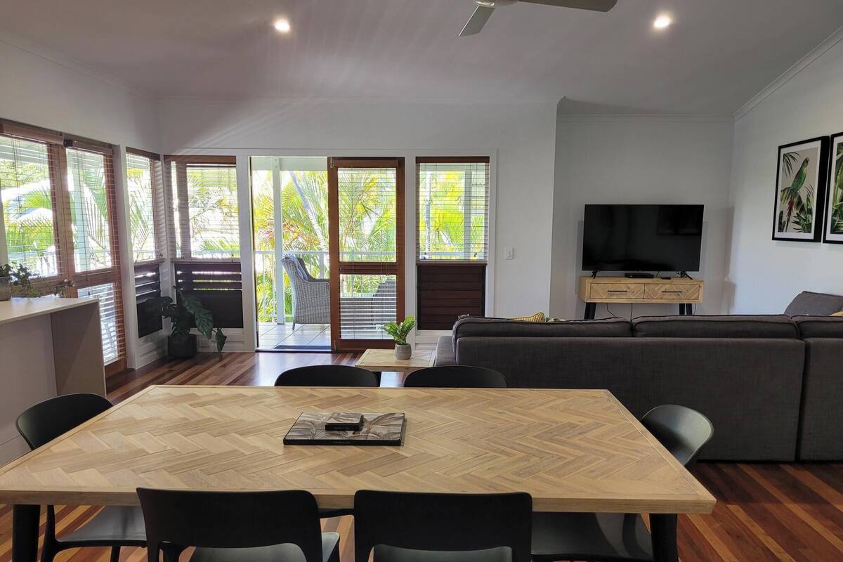 Una sala de estar moderna con suelo de madera en patrón de espiga, un sofá gris, un televisor sobre un mueble de madera y una zona de comedor con mesa de madera y sillas negras.