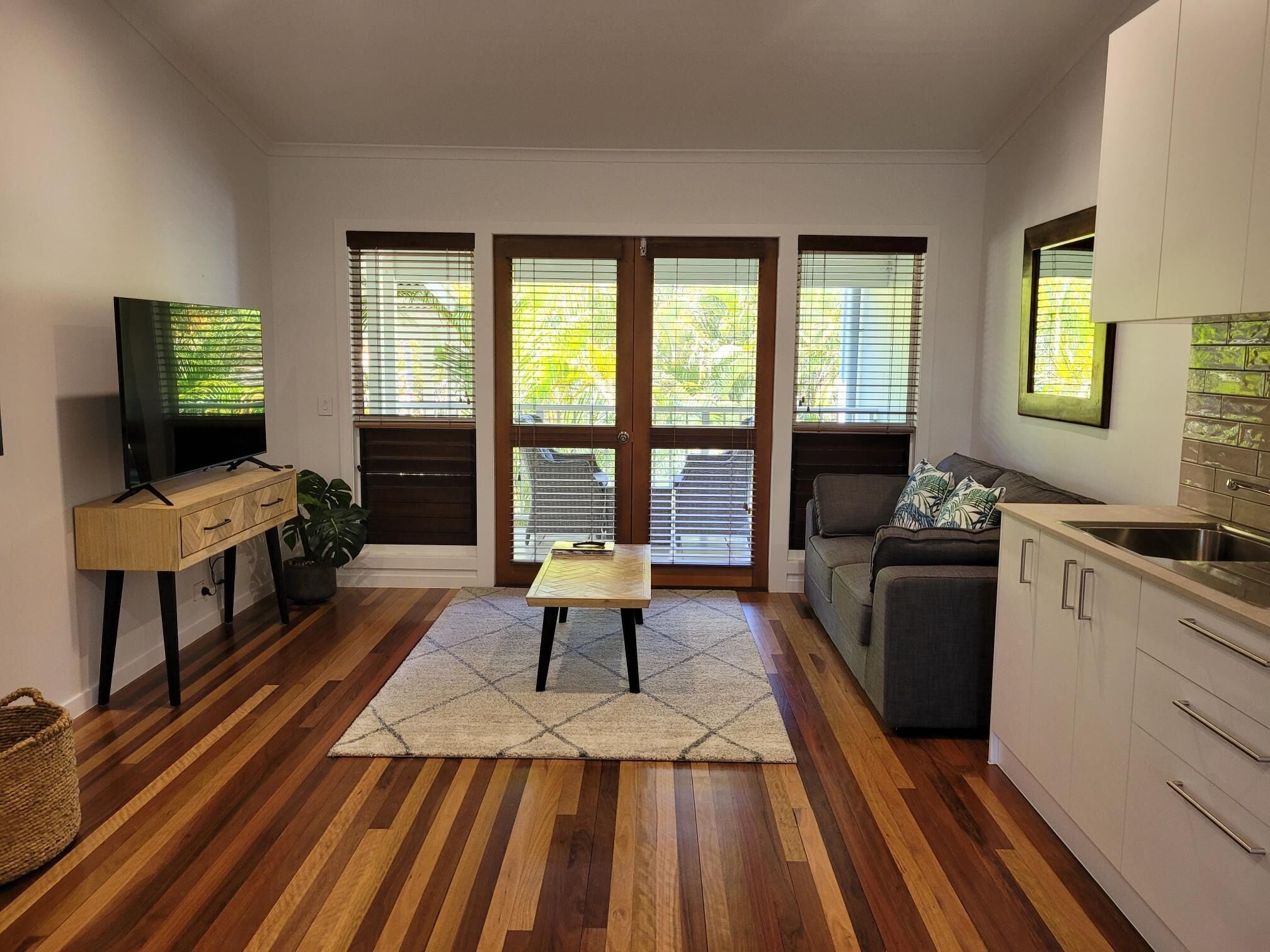 A compact living space with a wooden floor, a sofa, a TV, a small table, and a kitchen area with cabinets.