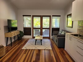 Un espacio habitable compacto con suelo de madera, un sofá, un televisor, una mesita y una zona de cocina con armarios.