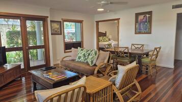 A spacious living room with a wooden floor, a sofa, wicker chairs, a dining table, and large windows with blinds.