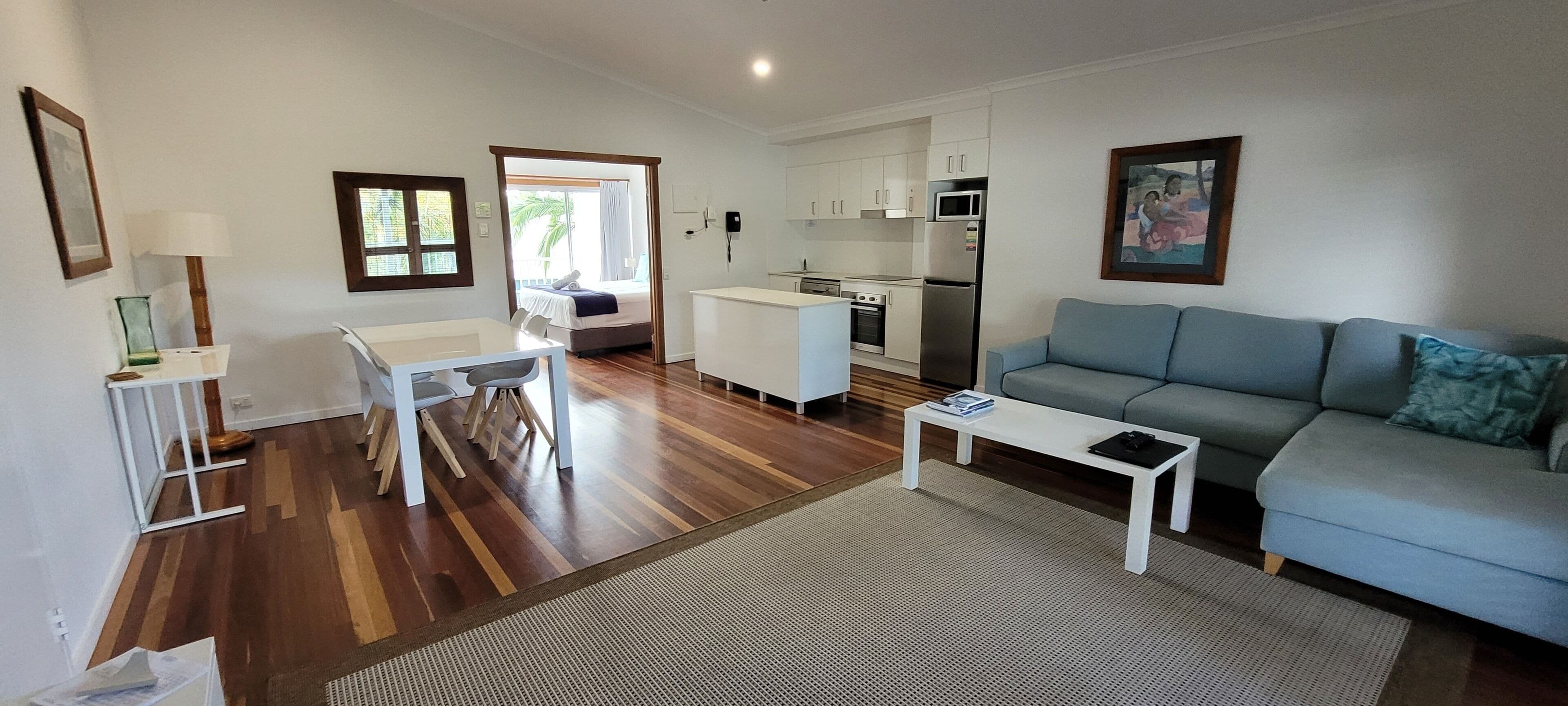 A modern living room with a sofa, coffee table, and dining area. There is a kitchen with white cabinets and appliances.