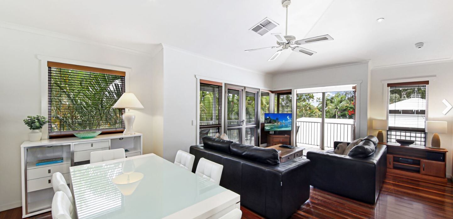 A modern living room with a glass dining table, black leather sofas, a ceiling fan, and large windows with blinds.