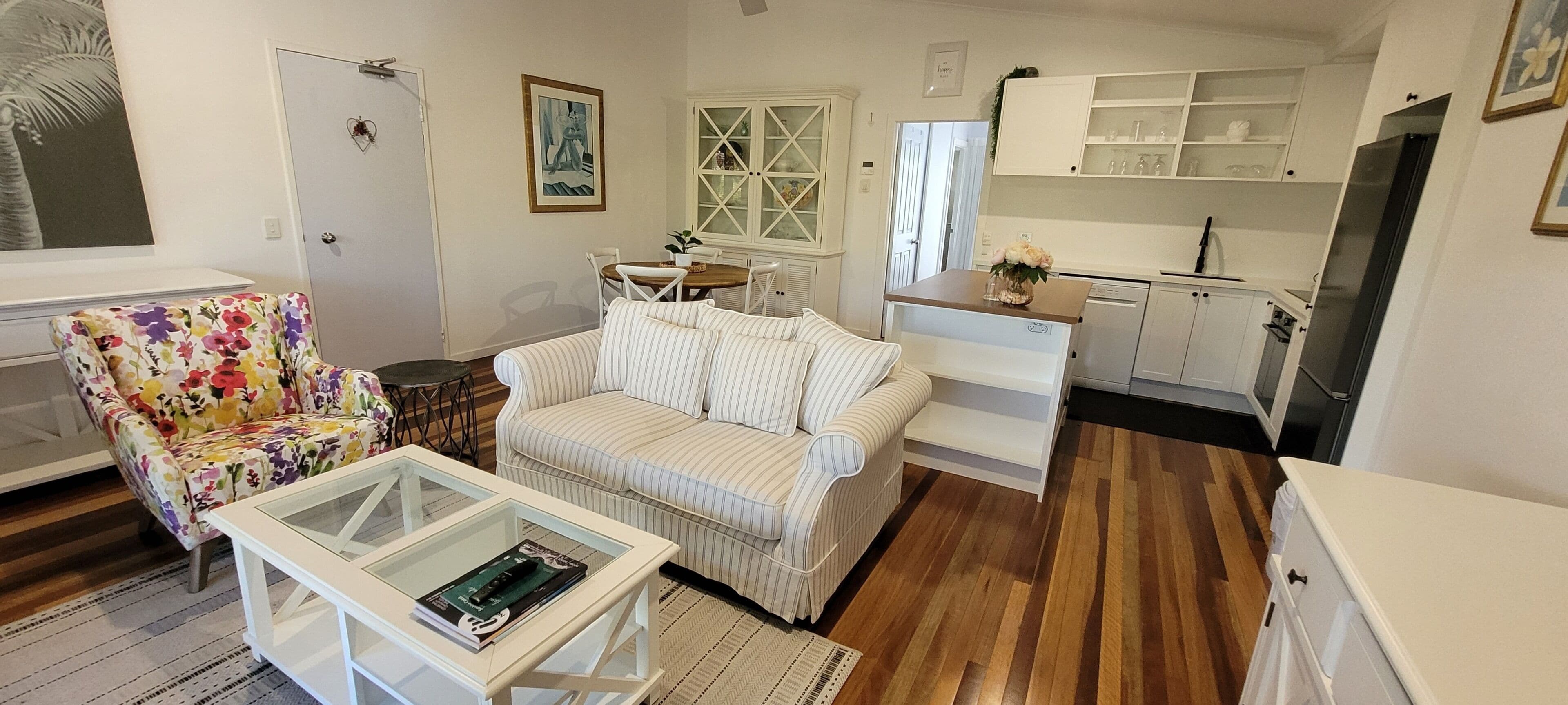 A compact living space with a floral armchair, striped sofa, and a glass coffee table. A kitchen area with white cabinets and a refrigerator is visible.