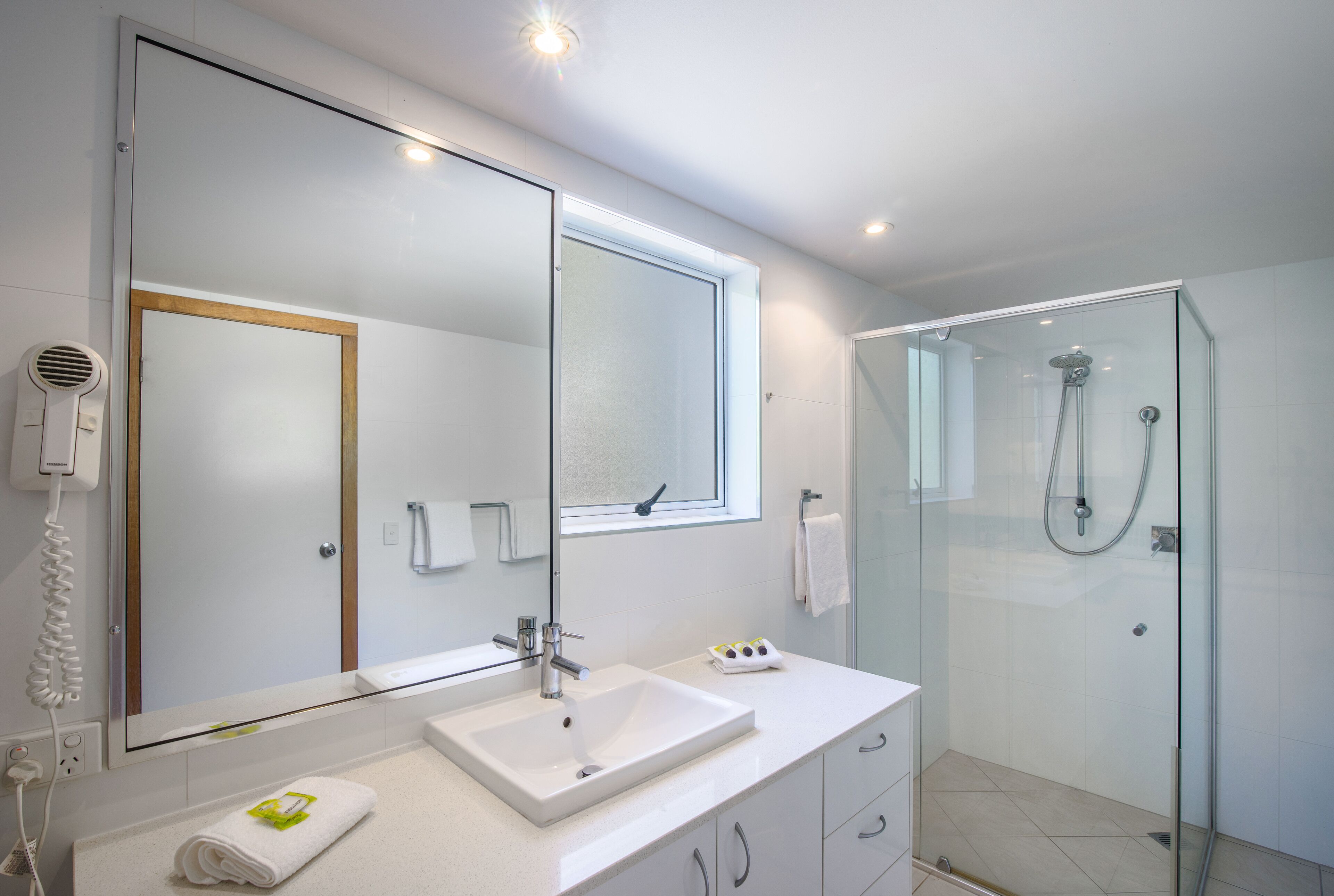 A modern bathroom with a large mirror, a white sink, a glass shower enclosure, and a hairdryer on the wall.