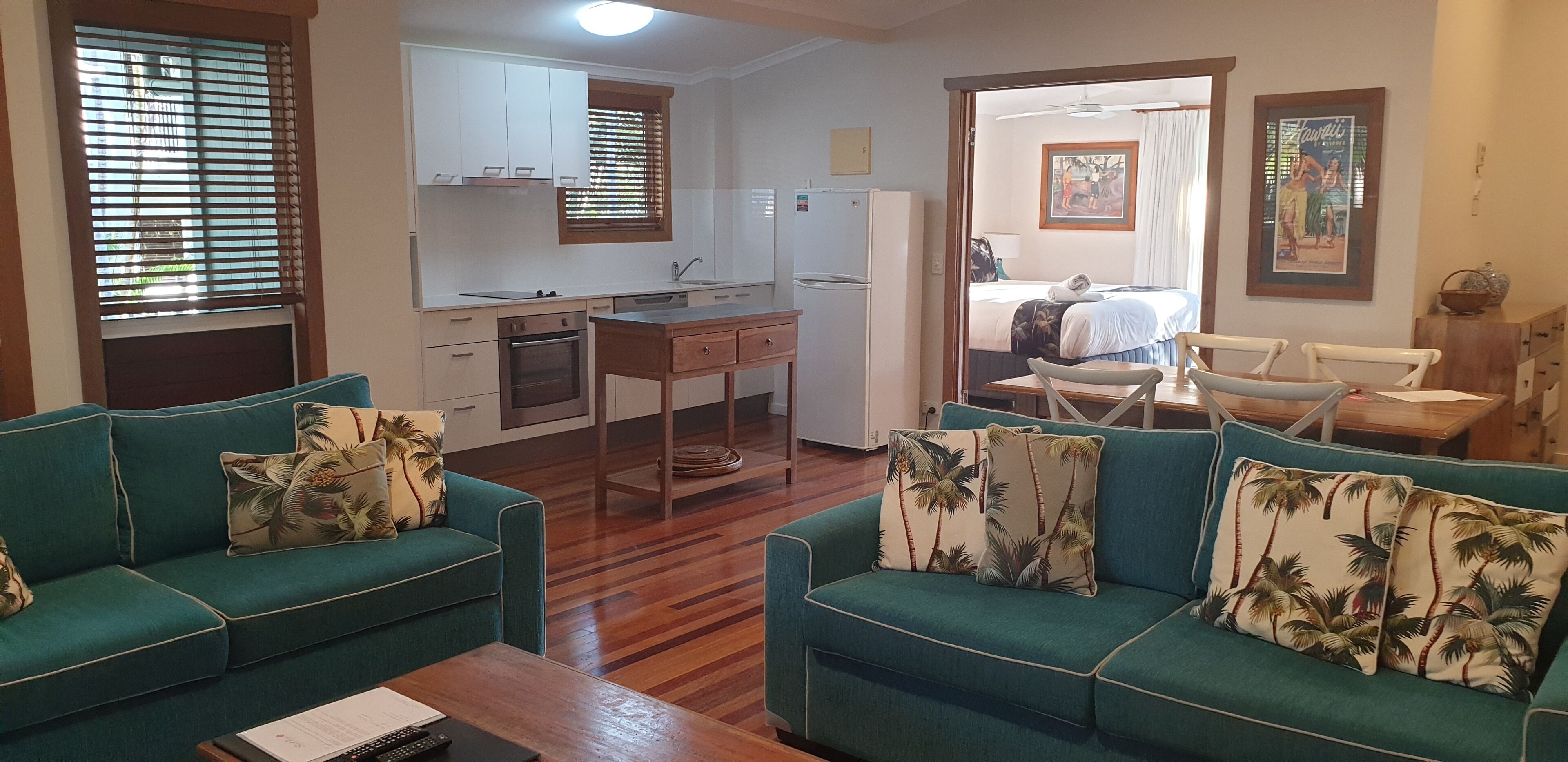 A compact living space with a kitchenette, two green sofas with tropical pillows, a dining table, and a bed in the background.