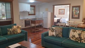 A compact living space with a kitchenette, two green sofas with tropical pillows, a dining table, and a bed in the background.