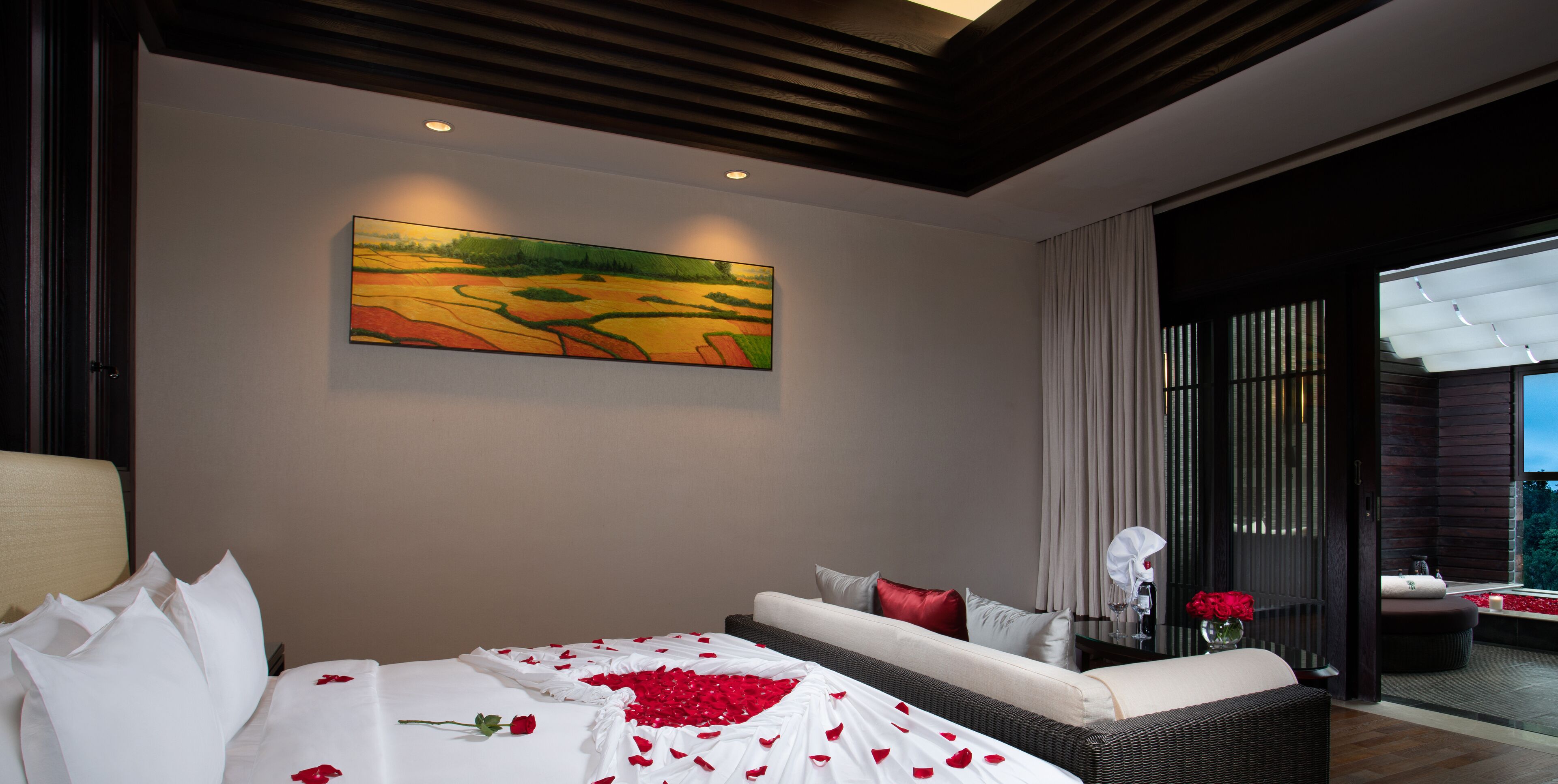 Habitación de hotel moderna con un cuadro grande, una cama con pétalos de rosa, un sofá y vistas a través de un ventanal.