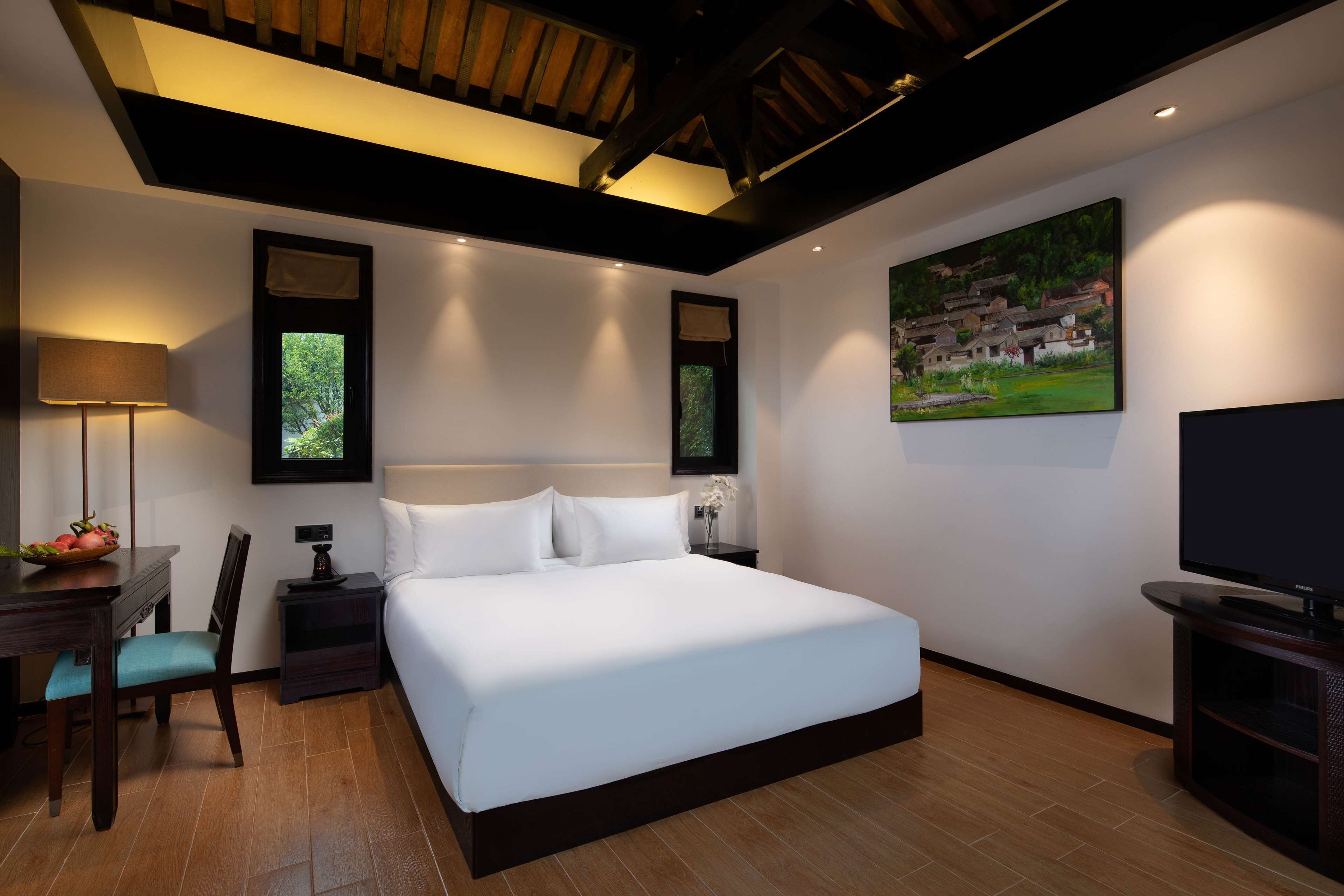 A modern hotel room with a large bed, a desk with a lamp, a chair, a TV, and a painting on the wall.