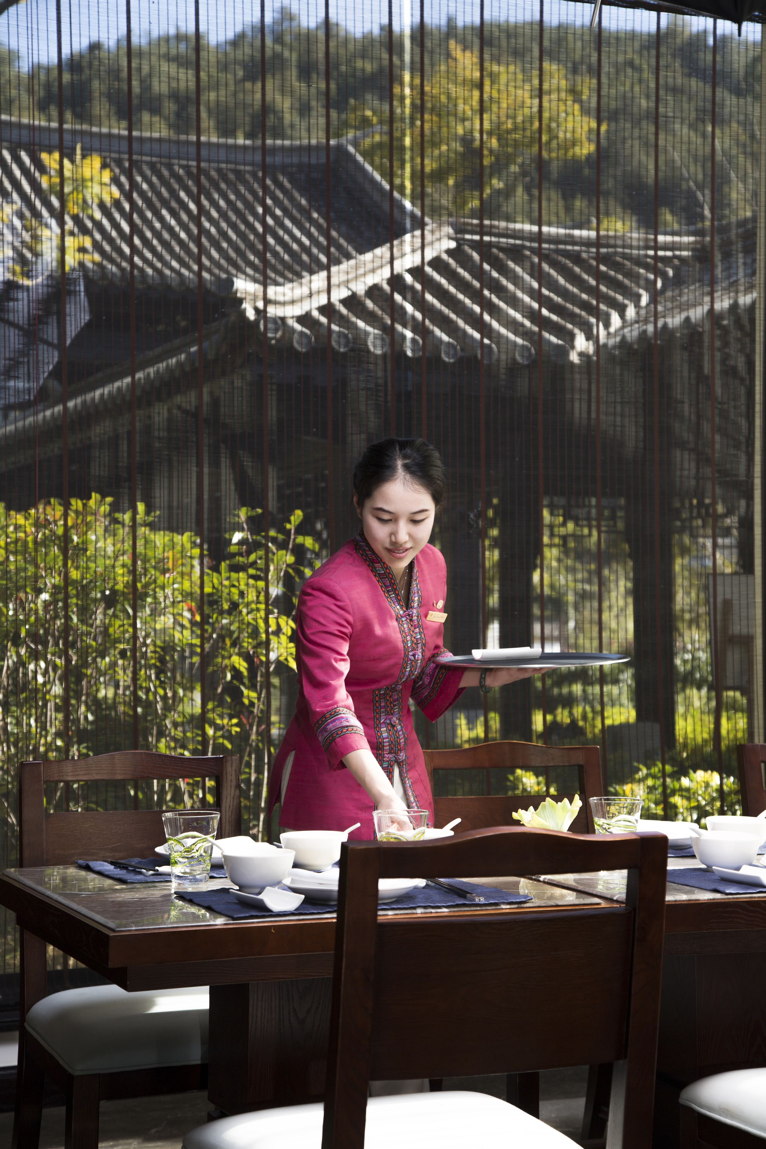 A dining area with a table set for two, a person serving food, and traditional architecture in the background.