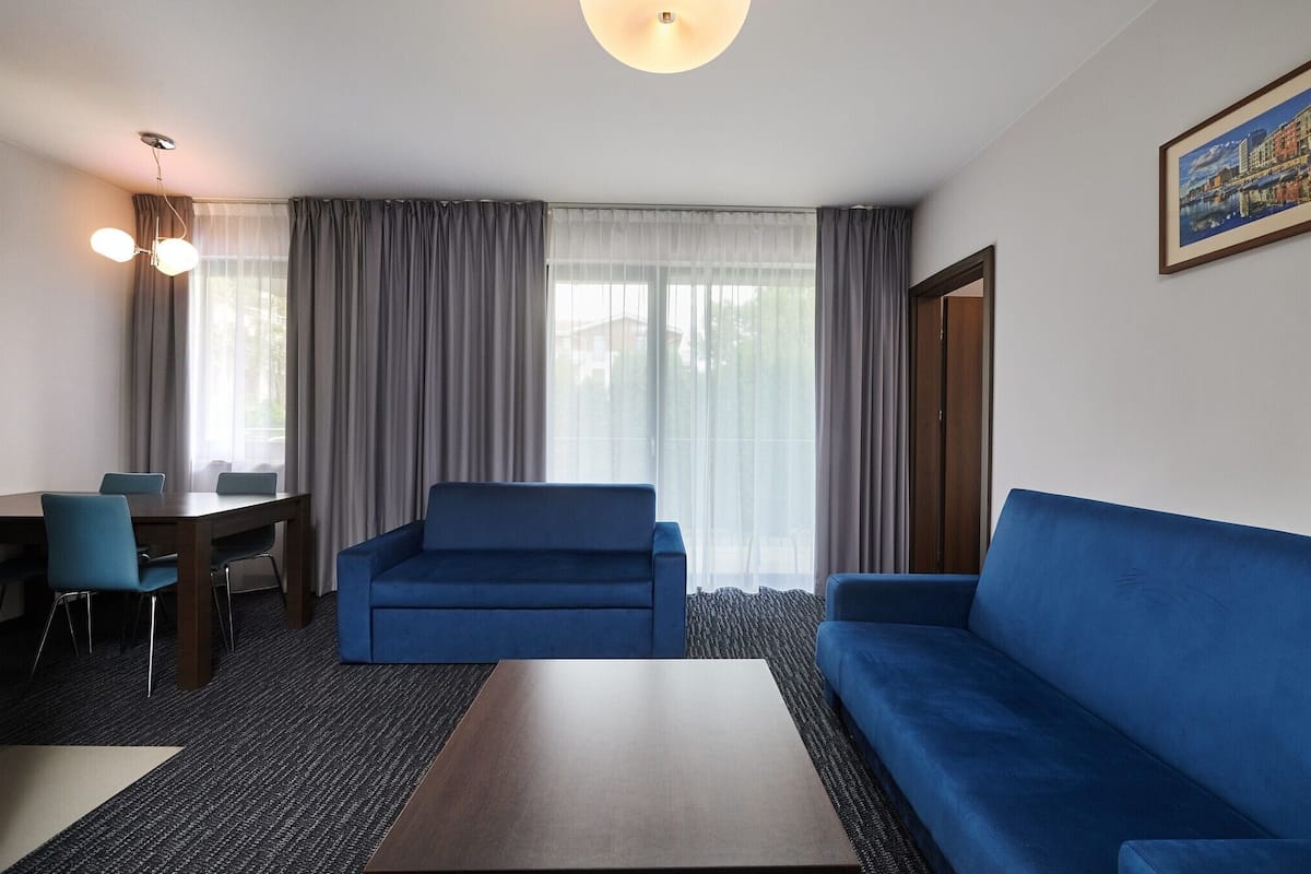 Ein modernes Hotelzimmer mit einem blauen Sofa, einem Holztisch, einem Esstisch mit Stühlen und einem großen Fenster mit Vorhängen.