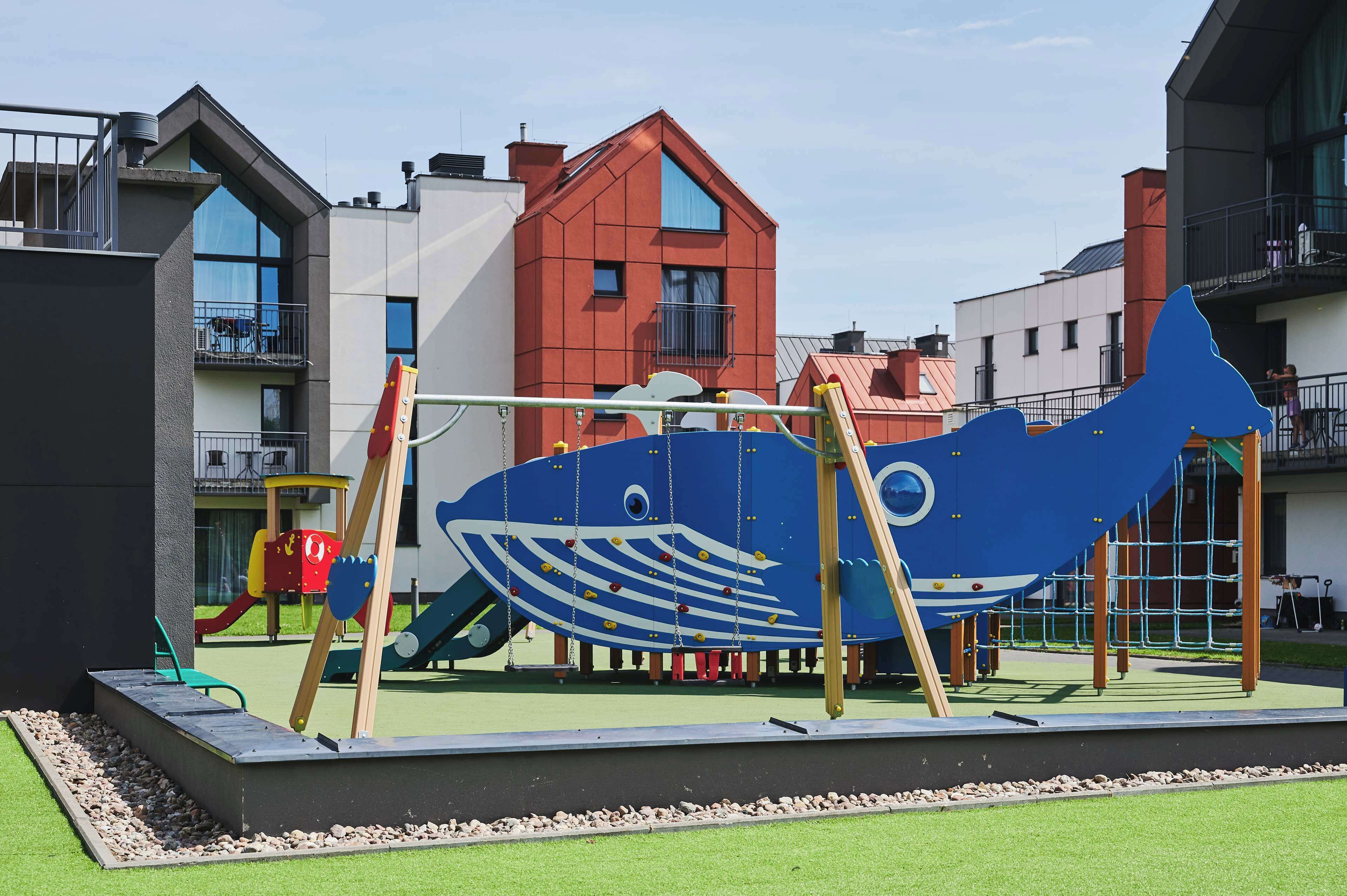 Un parque infantil con una gran estructura de escalada en forma de pez azul, rodeado de edificios.