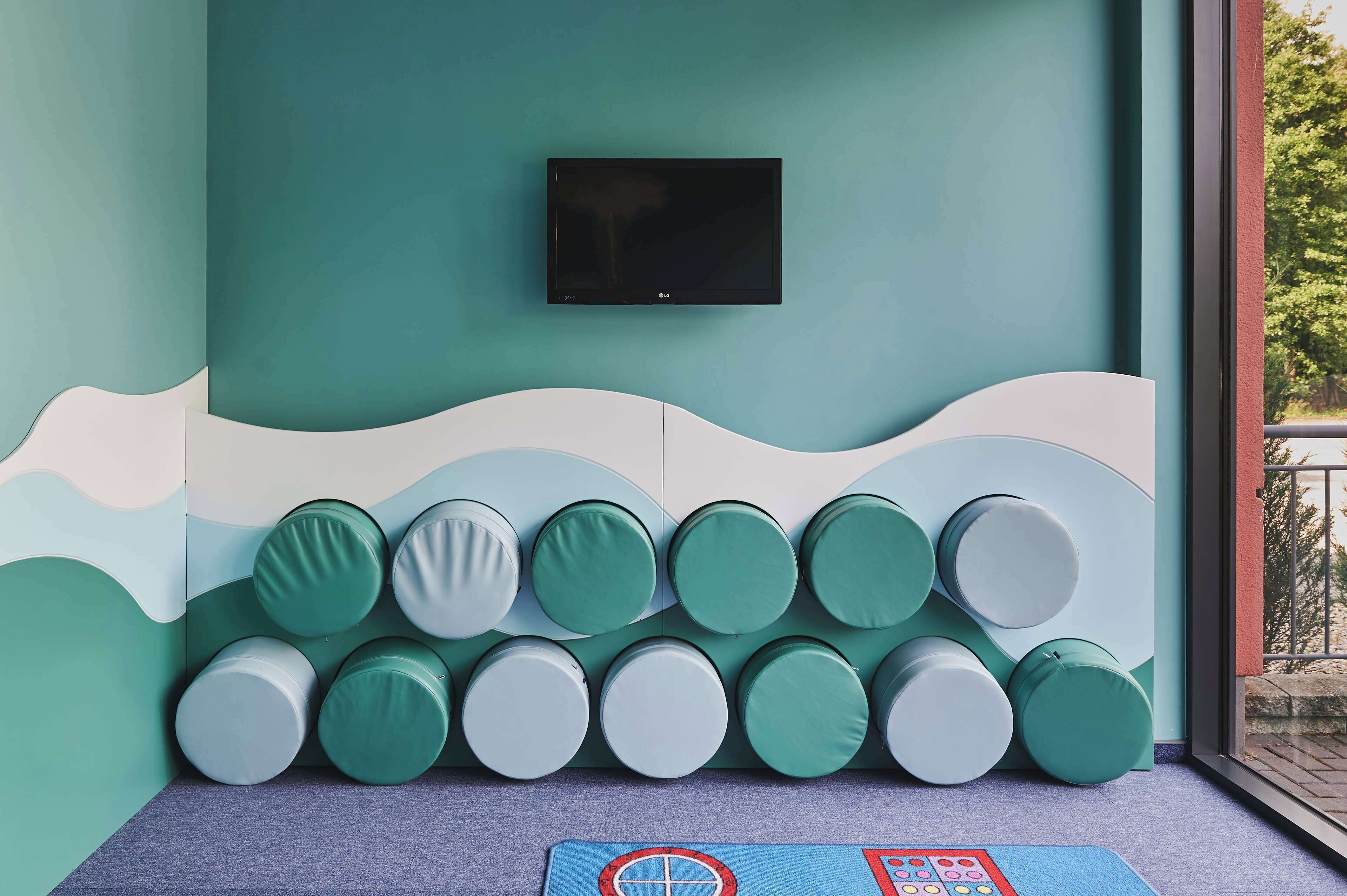 A room with a teal wall, a mounted TV, and a row of circular cushions against the wall.