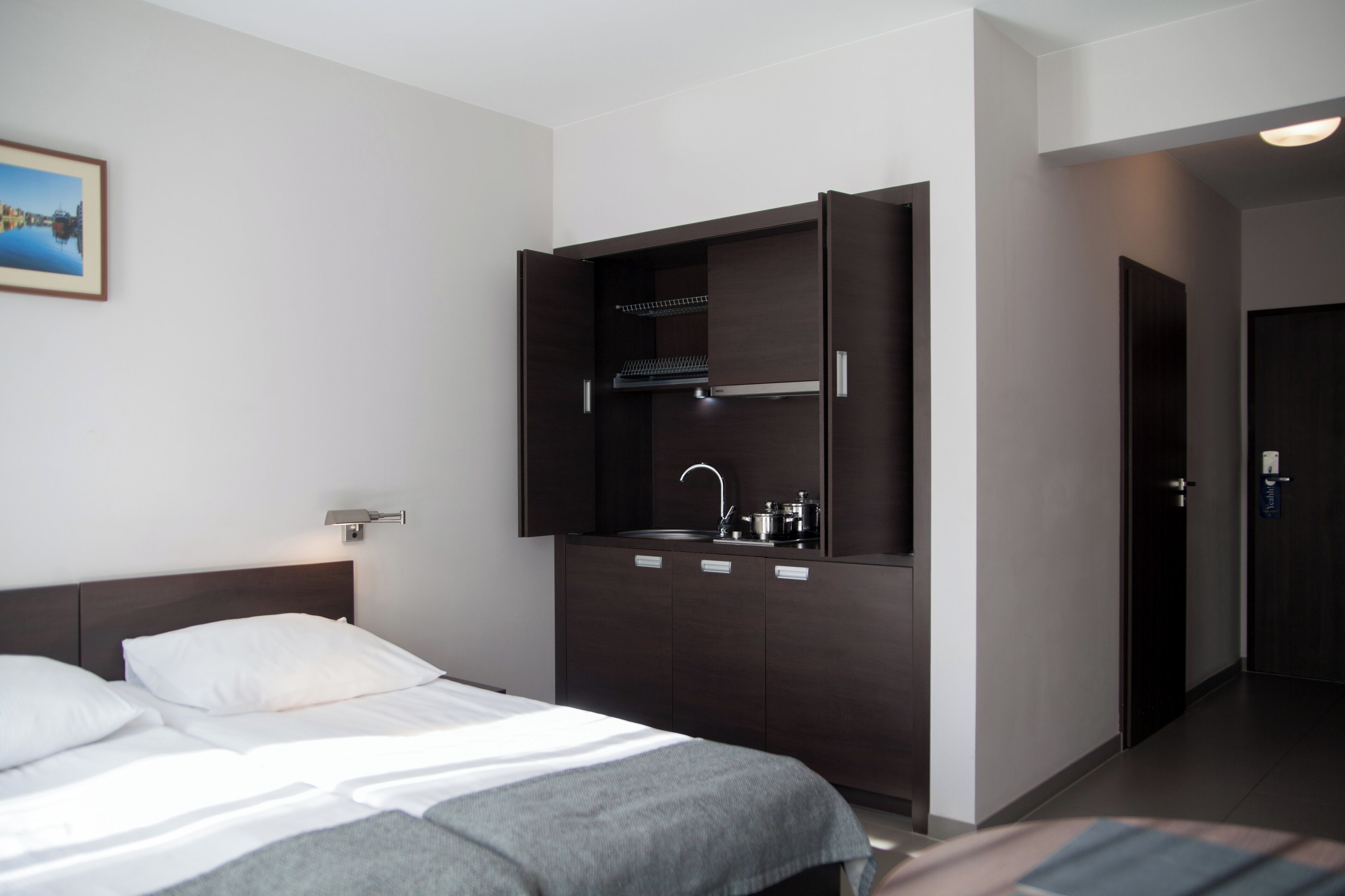 A modern hotel room with a bed, a built-in wardrobe, a small kitchenette, and a framed picture on the wall.