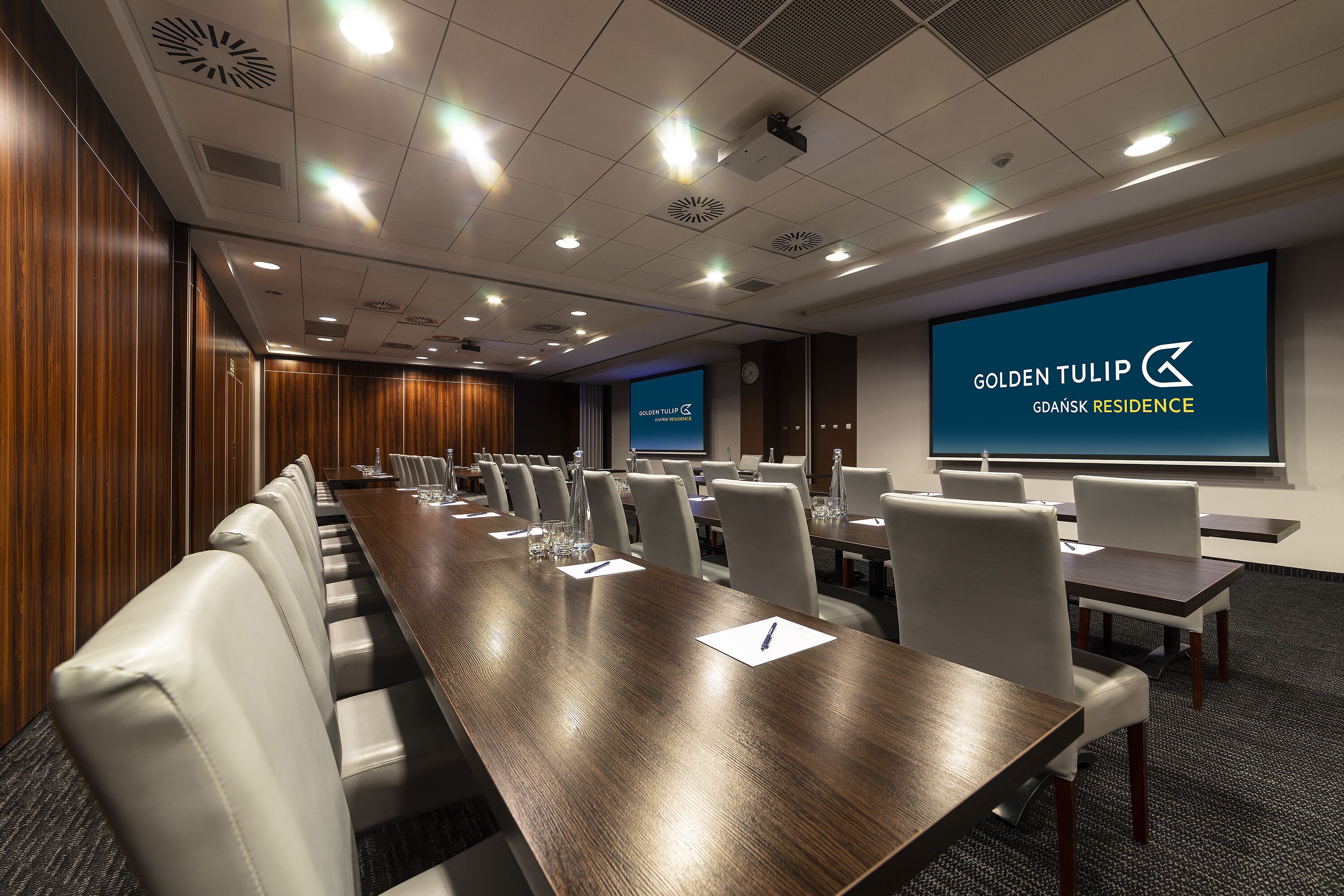 Uma sala de conferências com uma mesa comprida e várias cadeiras, uma tela grande exibindo "Golden Tulip Gdansk Residence" e um relógio na parede.