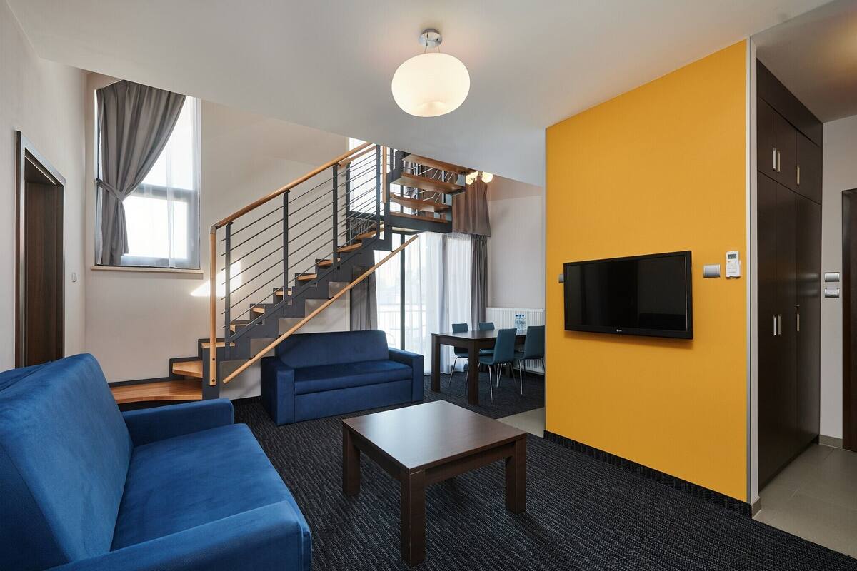 Ein modernes Wohnzimmer mit einem blauen Sofa, einem Holztisch, einer Treppe mit Metallgeländer, einem flachbildfernseher an der Wand und einer Wand in Gelb.
