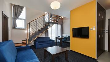 A modern living room with a blue sofa, a wooden coffee table, a staircase with a metal railing, a flat-screen TV mounted on the wall, and a yellow accent wall.