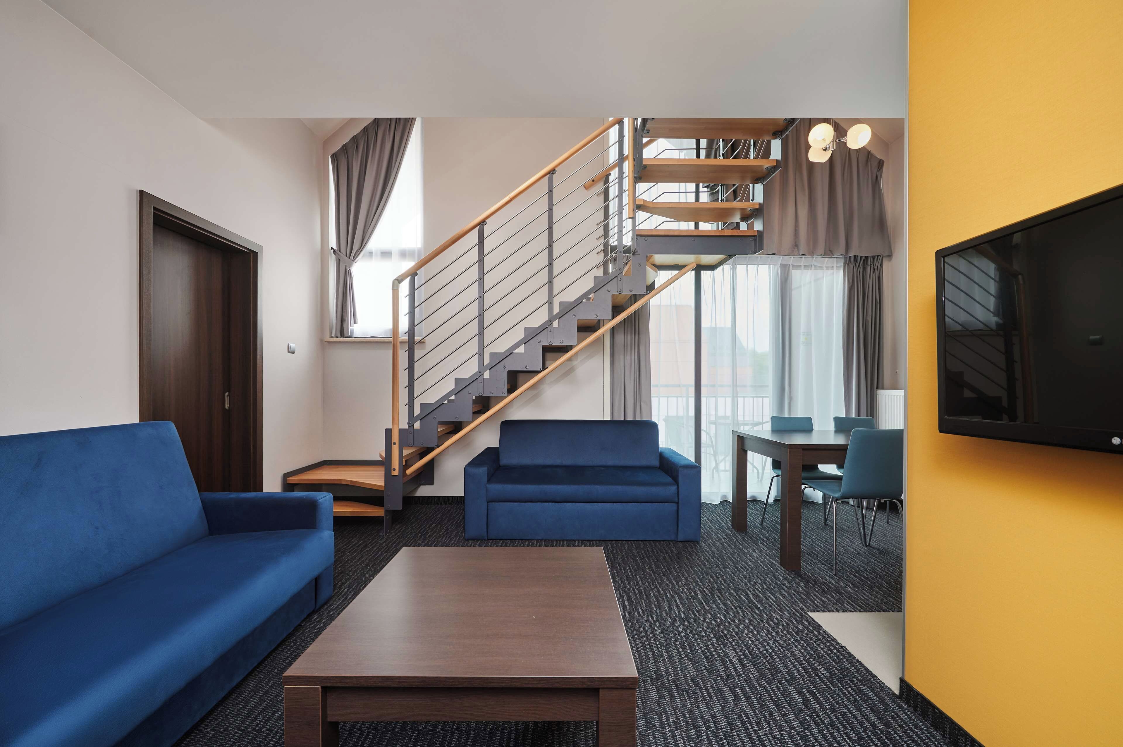 A hotel room with a wooden staircase, a blue sofa, a wooden coffee table, a TV, and a dining area with chairs.