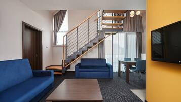 Quarto de hotel com um sofá azul, uma mesa de centro de madeira, uma televisão, uma escada e um espaço de jantar com mesa e cadeiras.