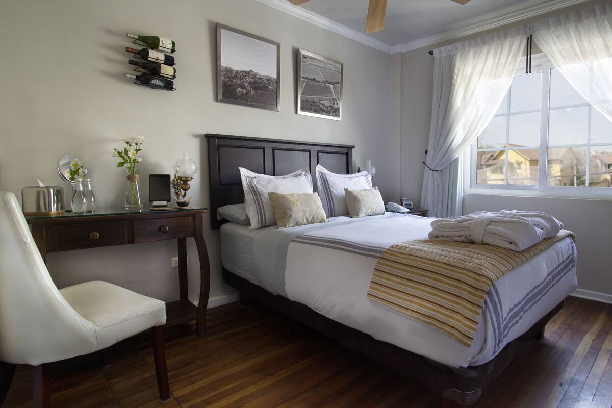 A bedroom with a bed, a desk, and a chair. There are two framed pictures on the wall and a window with curtains.