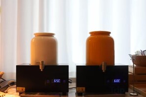 Two black digital clocks with numbers 6:51 and 6:01 displayed, placed on a table with two large ceramic containers, one beige and one orange, in front of a window with sheer curtains. - Milan Fashion Hotel (Shenzhen)