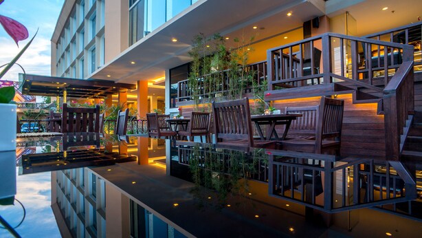 A modern outdoor seating area with wooden furniture and plants, reflecting on a glass floor. - Park Regis Kuta, Bali (Kuta)