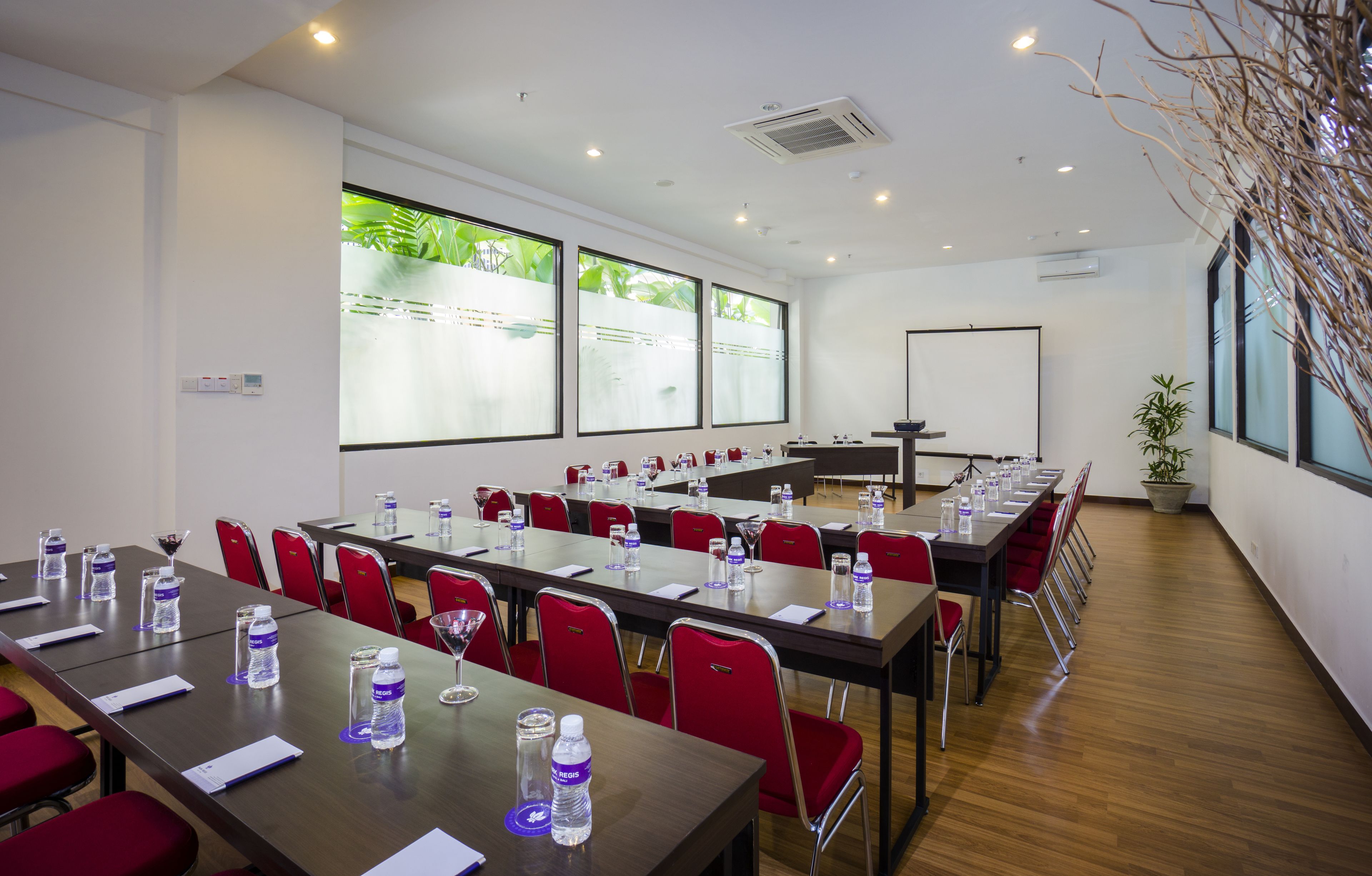 A conference room with multiple tables, red chairs, water bottles, and a projector screen.