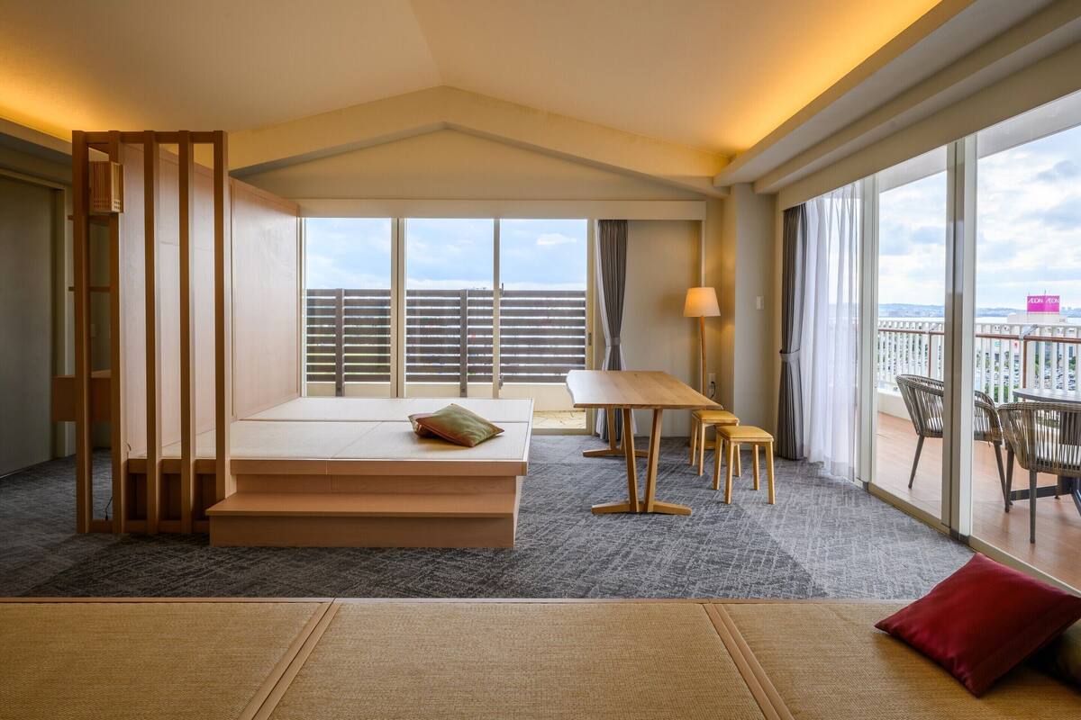 Ein Hotelzimmer mit großem Fenster, einem Holzbett, einem kleinen Tisch und einem Balkon mit Sitzgelegenheiten.