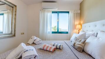 A hotel room with a bed, two rolled towels, a window with curtains, a bedside lamp, and an air conditioning unit.