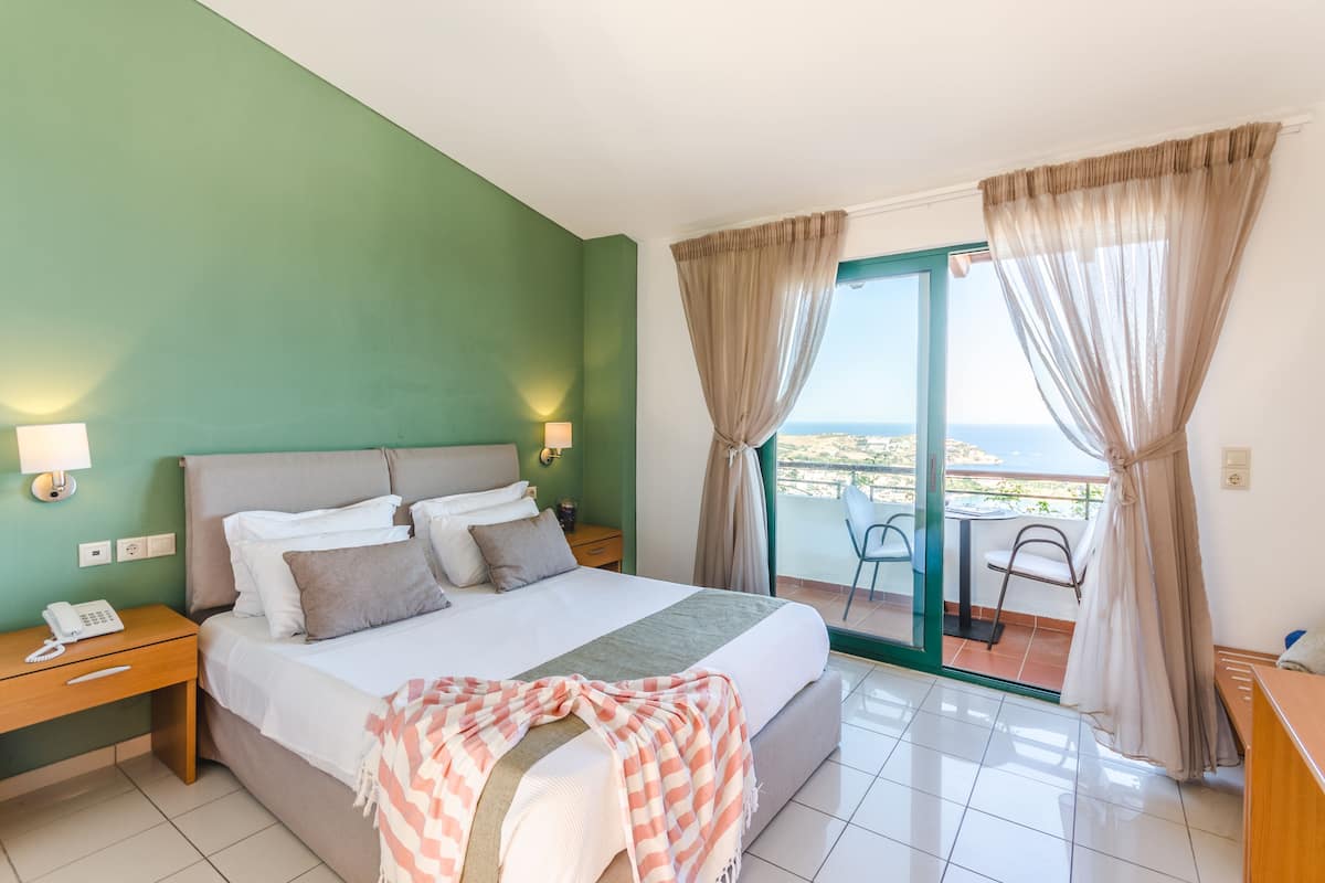 Une chambre d’hôtel avec un lit, des lampes de chevet, un téléphone, un balcon avec une chaise et une vue sur la mer.