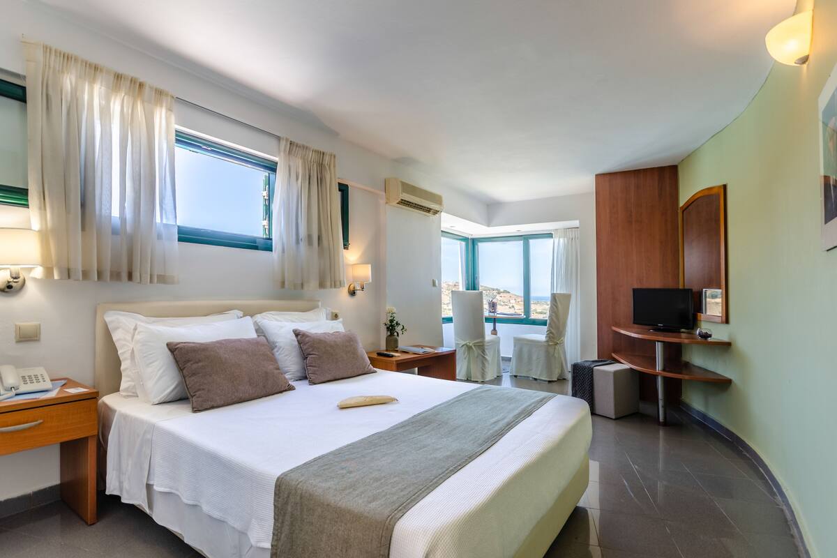 Une chambre d’hôtel avec un grand lit, un bureau avec un téléphone, un téléviseur posé sur un meuble en bois et une fenêtre avec des rideaux.