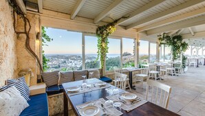 An outdoor dining area with tables set for two, large windows offering a view of the city, and a covered roof structure. - Pela Mare Hotel (Malevizi)