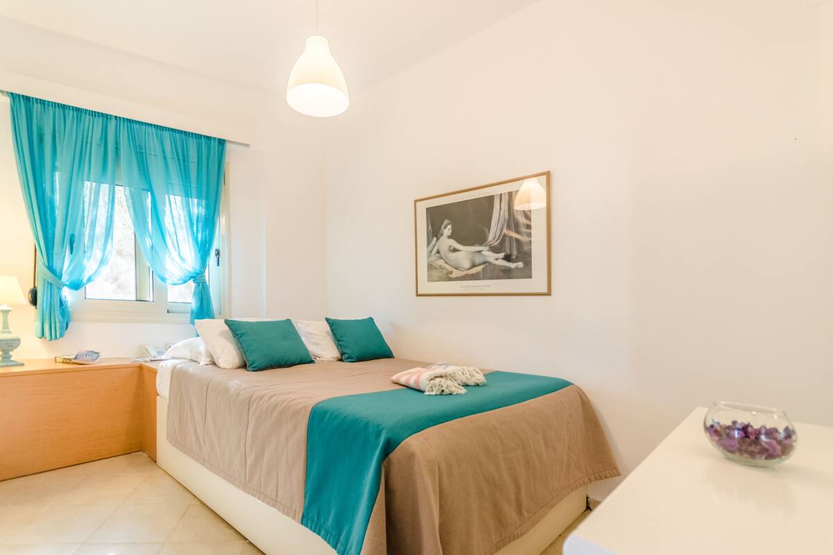 Une chambre à coucher avec un lit, une table de chevet, une lampe, une fenêtre avec des rideaux, un tableau encadré au mur et une petite table avec un bol.