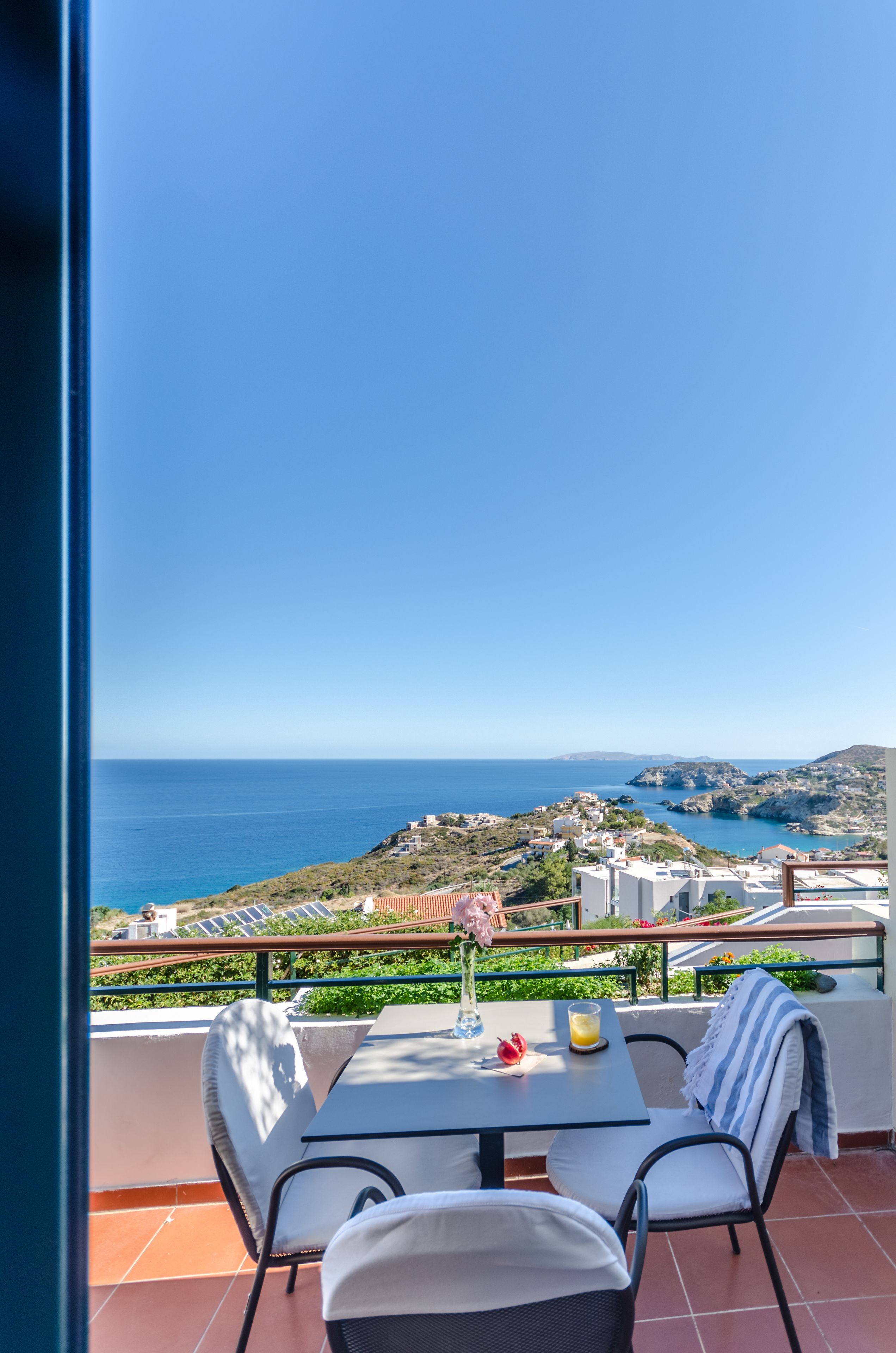 Un balcón con mesa y sillas, con vista a un paisaje costero con edificios y el mar.