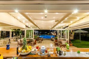 Uma área de refeições ao ar livre com vista para a piscina, mesas e cadeiras de madeira, e diversos alimentos e bebidas sobre as mesas.