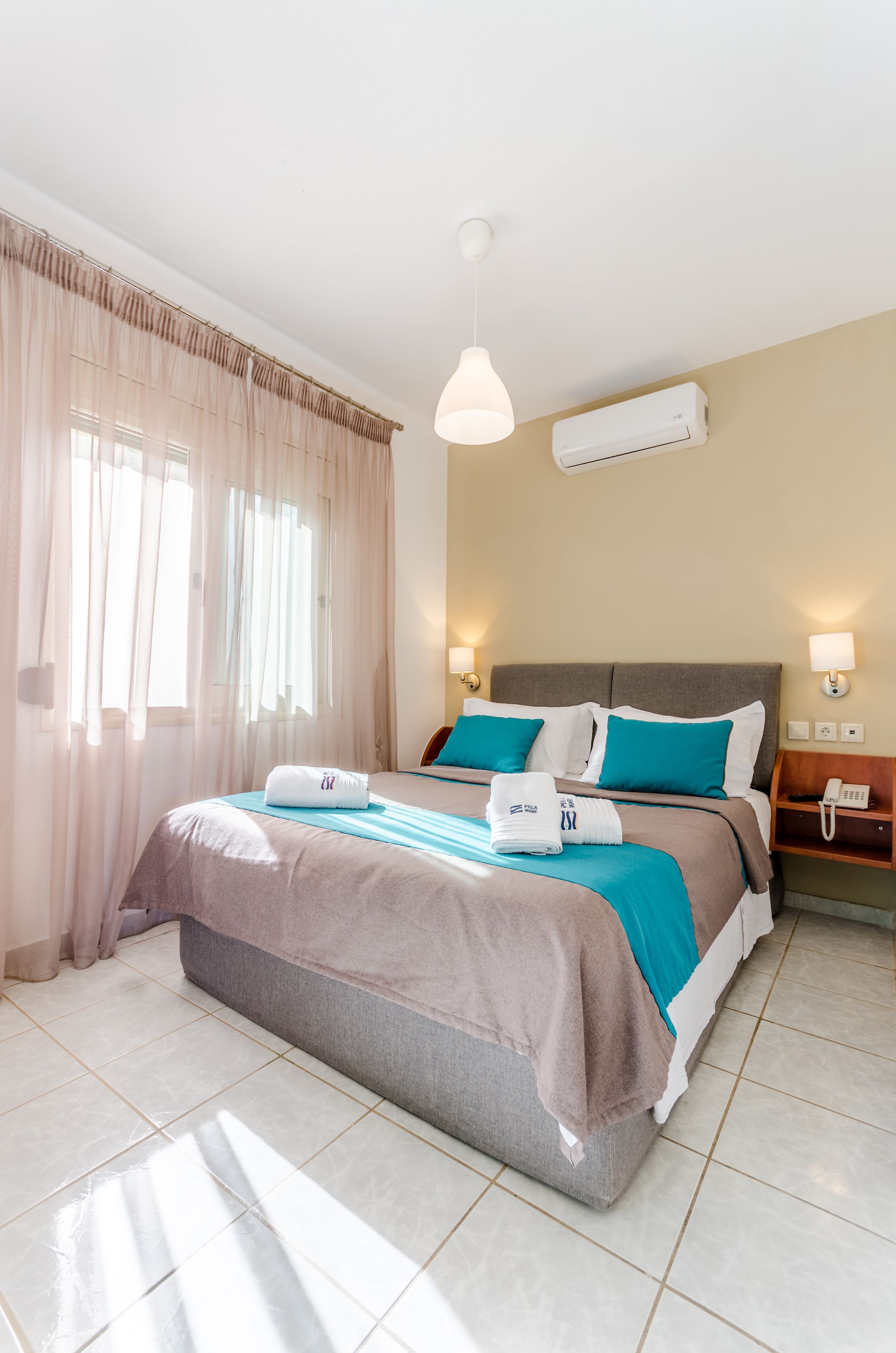 A hotel room with a bed, curtains, an air conditioner, a bedside lamp, and a telephone on a nightstand.