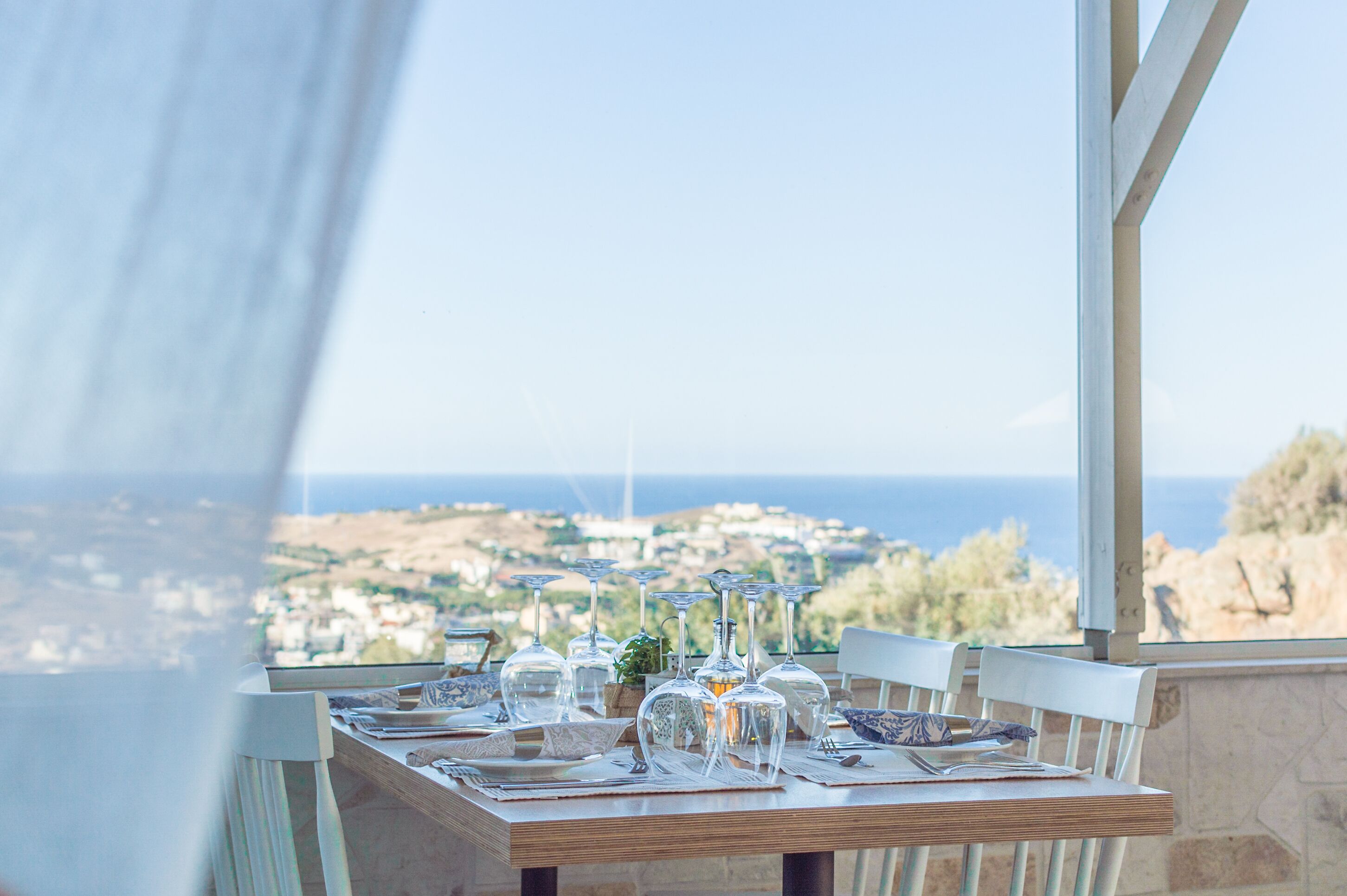 A table set for dining with wine glasses, plates, and cutlery, overlooking a coastal town.