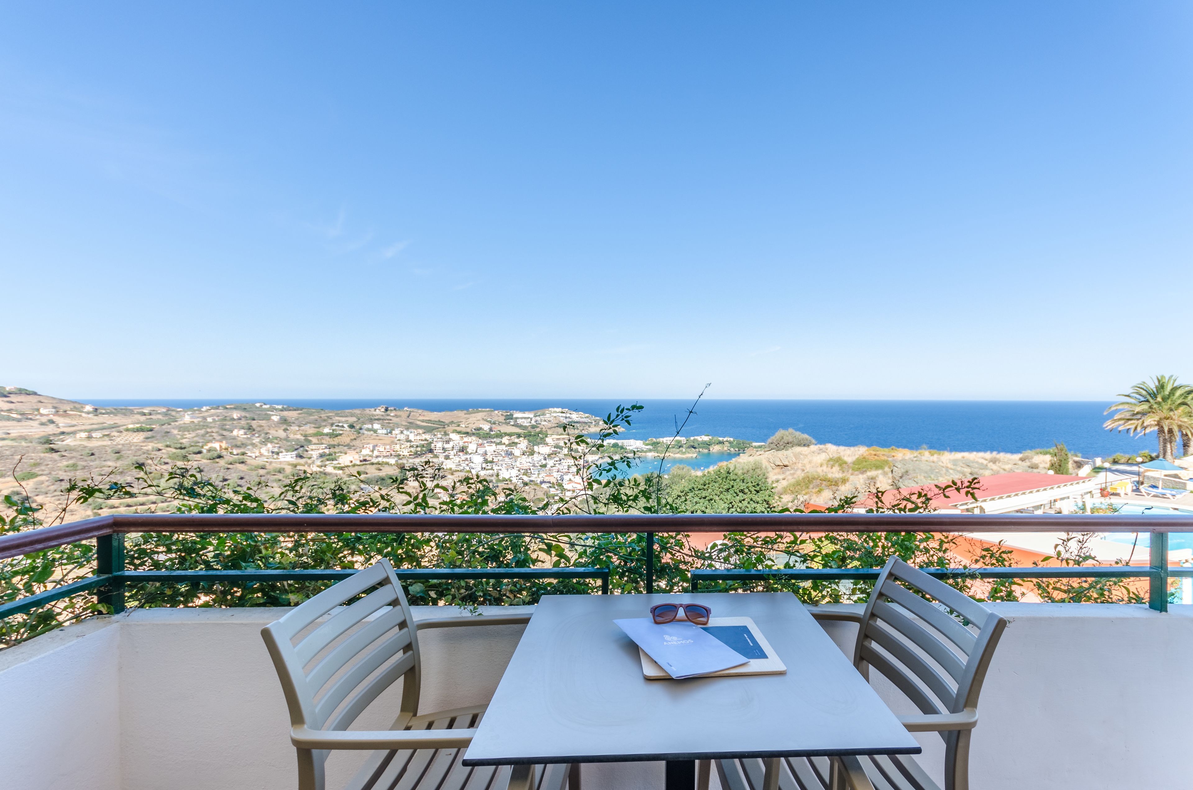 A balcony with a table and chairs, overlooking a coastal town and the sea.