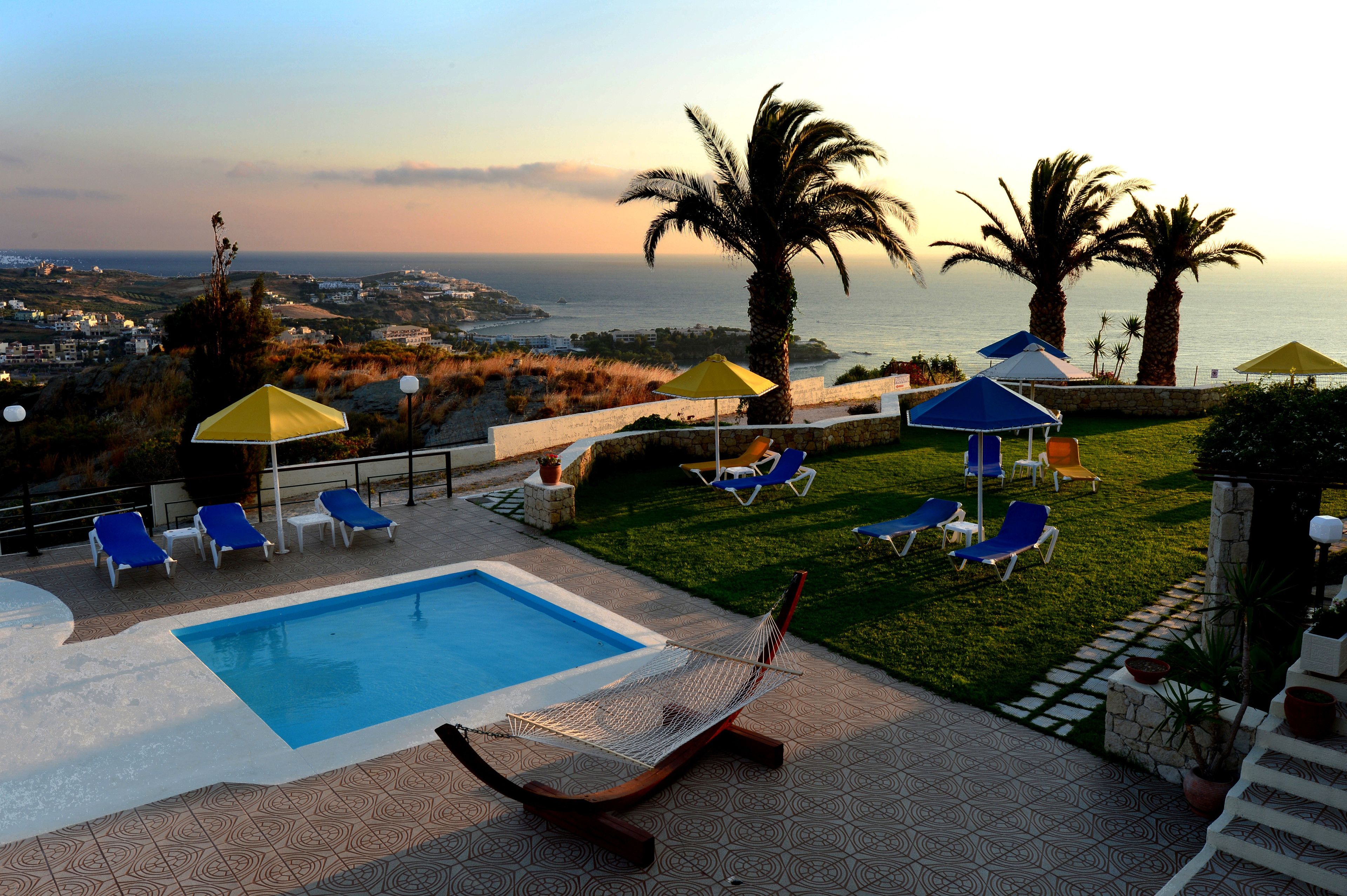 Un área de piscina con sillas de descanso y sombrillas, una hamaca y vistas al mar y al paisaje distante.